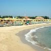 Spiaggia sabbiosa con mare calmo, lettini e ombrelloni gialli, capanne in lontananza.