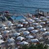 Spiaggia affollata con ombrelloni bianchi, sedie a sdraio, piscina e mare.